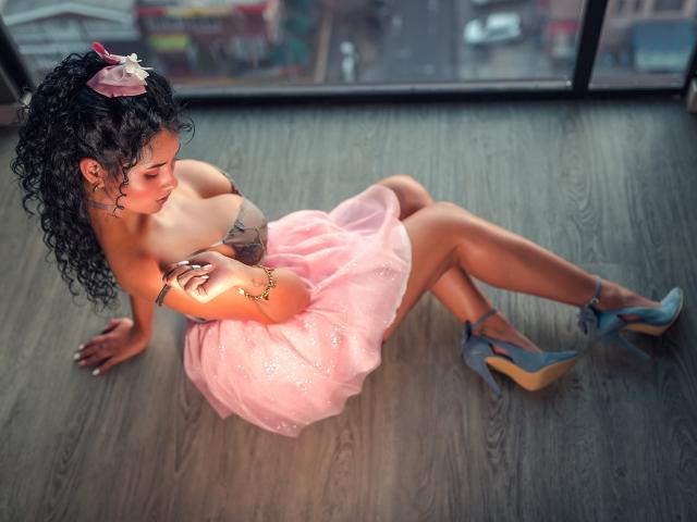 CanelaLebrad with curly black hair, pink tulle dress, and blue heels, sitting on wooden floor, showing her tits, tattoo on left shoulder, bracelet on right wrist, looking down.