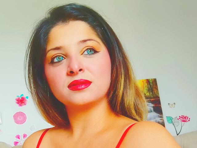 ScarletOra has bright blue eyes, shiny red lipstick, and brown hair with blonde highlights. She's wearing a red strap top, and has bold, winged eyeliner.