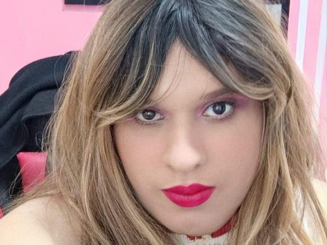 KendricHot tem cabelo loiro escuro com mechas pretas, sombra rosa e batom vermelho vibrante. Eles estão olhando diretamente para a câmera com uma expressão séria..