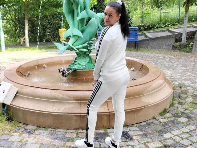 JessieStar69 with black ponytail, white tracksuit, and black-and-white sneakers, standing by a circular fountain with a green statue, looking back with a slight smile.