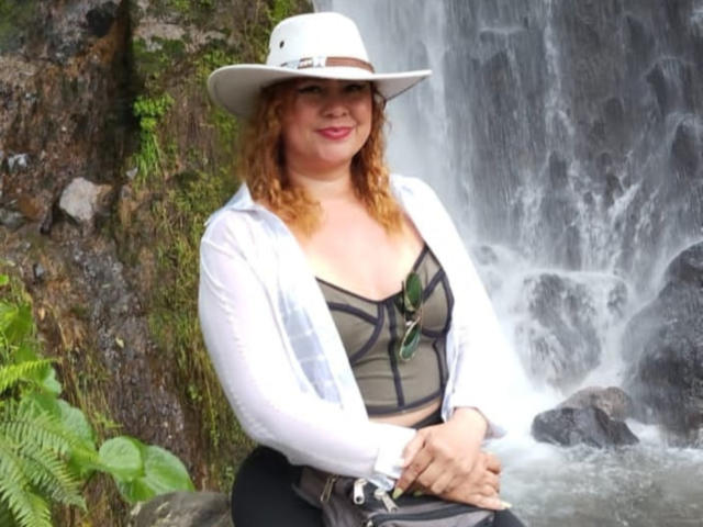 Madamecollinss with curly red hair, wearing a white hat, sheer white jacket, black bra, and black pants, standing by a waterfall, holding sunglasses in her hands.