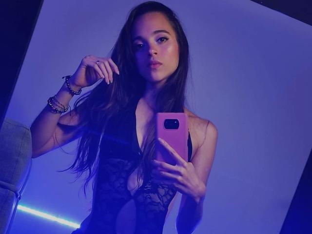 VioletaHade with long brown hair, wearing a black lace bodysuit, holding a pink phone, blue lighting, bracelets on her right wrist, looking at the camera.