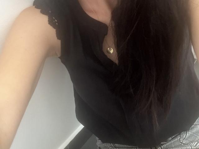 LanyJolene con cabello negro largo, un top negro sin mangas con encaje, un collar de botones dorados y pantalones grises a cuadros, mostrando su brazo y pecho desnudos. Los senos son parcialmente visibles..