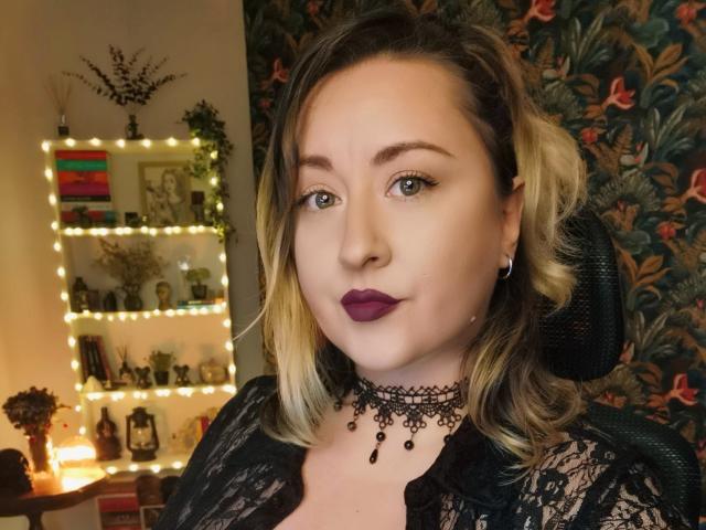 LuxNoir has blonde hair, purple lipstick, black lace choker, and black lace top, sitting in a chair with a black headrest. Her tits are medium, and she's looking at the camera.