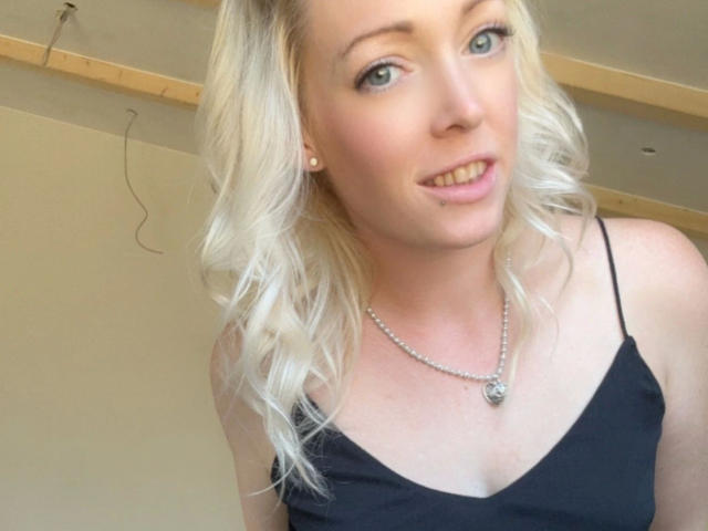 LeaKirsch-hot has blonde hair, blue eyes, and a cute smile. She's wearing a black spaghetti strap top and a silver necklace with a pendant. Boobs visible, tits noticeable.