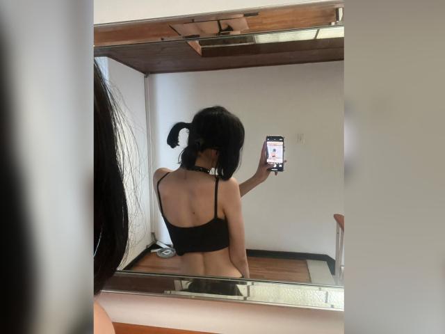 MillaHottLove69 with black pigtails, back to mirror, wearing black strapless top, exposing her bare ass, taking a selfie with her phone, choker around her neck, wood floor.