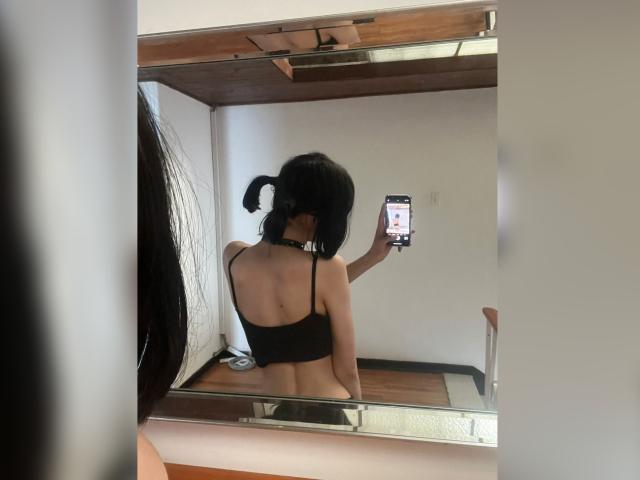 MillaHottLove69 mit schwarzen Zöpfen, mit dem Rücken zum Spiegel, macht ein Selfie mit ihrem Handy, trägt einen schwarzen BH und zeigt ihren Hintern, in einem Raum mit weißen Wänden und einer Holzbalkendecke..