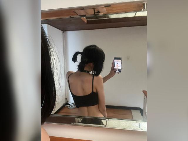 MillaHottLove69, com maria-chiquinhas pretas, sutiã preto e colar, tirando uma selfie em um espelho, mostrando suas costas e nádegas nuas, segurando um celular..
