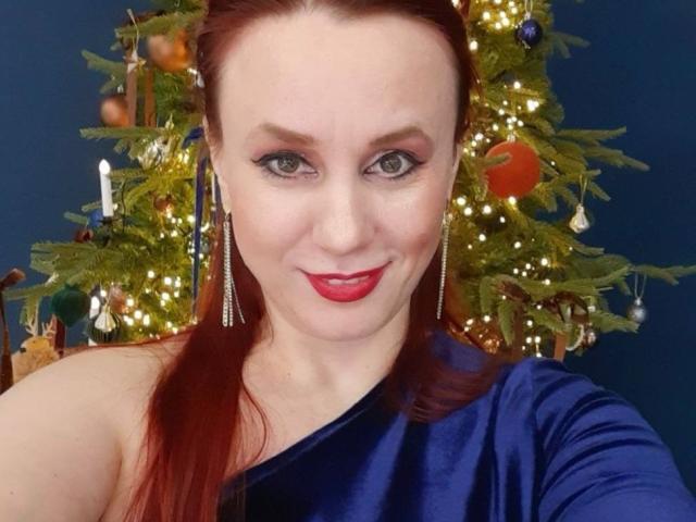 KristiVegas has red lipstick, blue dress, and long red hair, standing in front of a decorated Christmas tree with lights and ornaments, smiling at the camera.