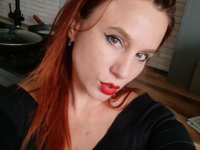KristiVegas with red lipstick, black eyeliner, and red hair, looking at the camera with a slight pout, wearing a black top, in a kitchen. Her tits are small.