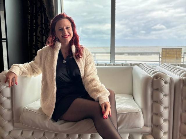 KristiVegas with red hair, smiling, wearing a fluffy white cardigan, black dress, and black tights, sitting on a white tufted sofa, legs crossed, red nails, necklace.