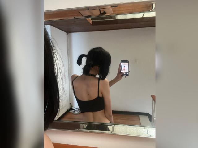 MillaHottLove69 with black pigtails, black bra, and choker, taking a selfie in a mirror, showing her bare back and ass. Holding a phone, reflecting in a wooden-framed mirror.