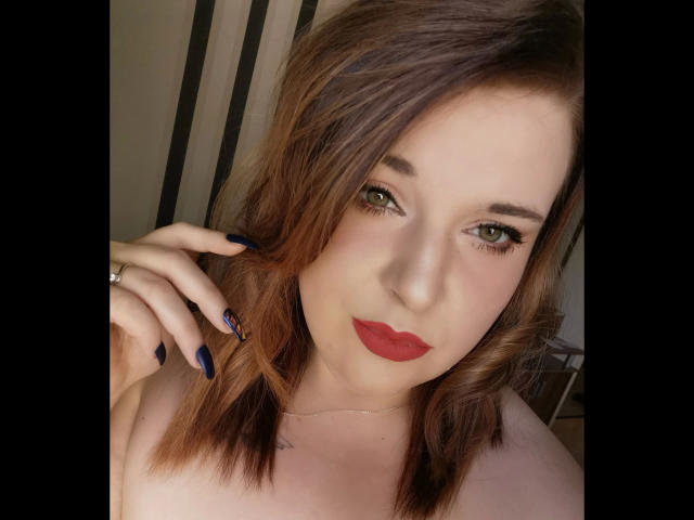 CashladyJacky97-hot with red lips, green eyes, and dark blue nails, touching her brown, wavy hair, wearing a silver ring on her left hand, and looking at the camera.