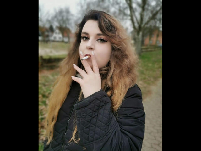 CashladyJacky97-hot with long, wavy brown hair, smoking a cigarette, wearing a black quilted jacket, has a pensive expression, standing on a gravel path, overcast day.