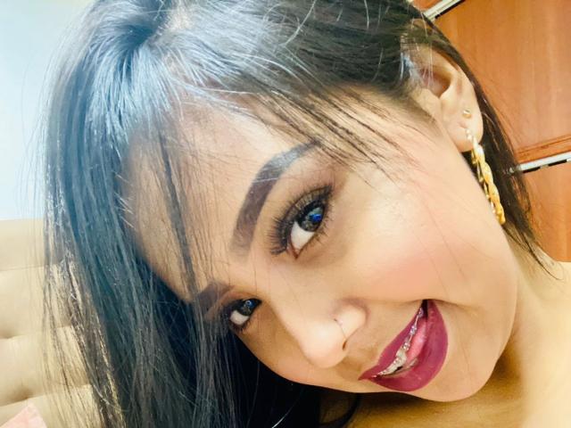 AliciaRichie's face, dark hair, brown eyes, thick eyebrows, red lipstick, smiling, gold chain earrings, light skin, nose piercing, slight tilt to head, focused on camera.