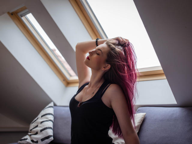 EmmaLust with pink-tipped hair, black tank top, arm raised, hand in hair, eyes closed, red lipstick, sitting on grey sofa, sunlight from slanted windows, relaxed, confident, sexy.