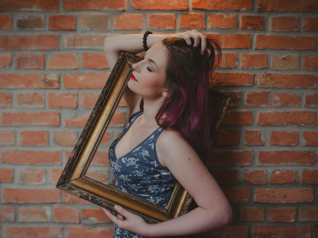 EmmaLust with purple-dyed hair, holding a gold-framed mirror against a brick wall, wearing a blue floral dress, arms above her head, red lipstick, and a black bracelet.