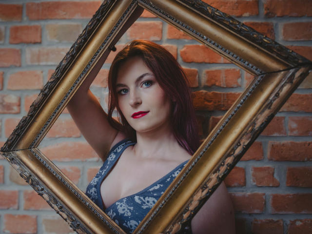 EmmaLust with red lipstick, blue floral dress, and arm in gold frame, looking at camera, showing her tits, against brick wall. She's got purple hair and a sexy smile.