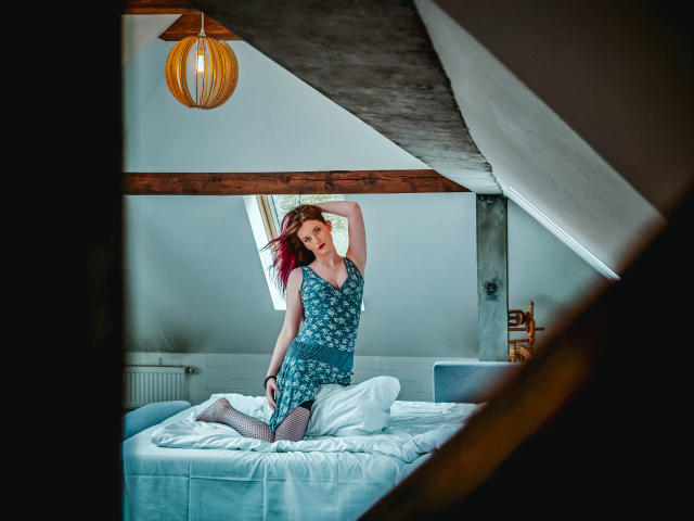 EmmaLust with red hair, wearing a blue floral dress, kneeling on a bed, arms raised, showing her tits, in a room with a wooden ceiling and a hanging lamp.