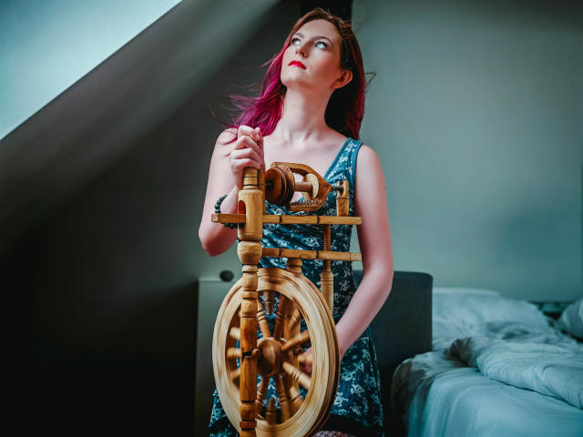 EmmaLust with pink hair, red lipstick, holding a wooden ship's wheel, wearing a blue floral dress, standing in a dimly lit room, looking up with a slightly curious expression.