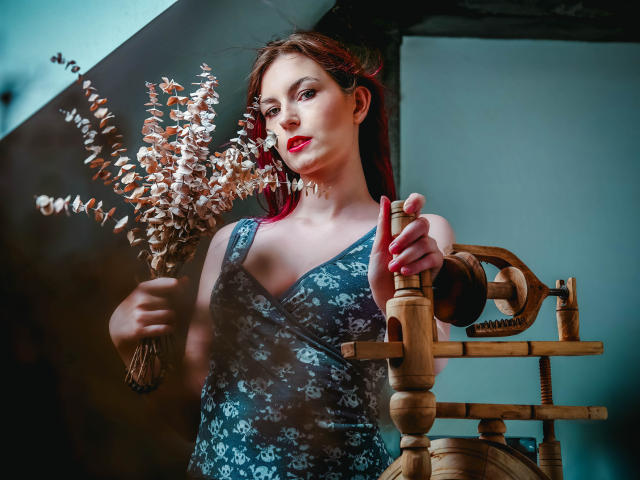 EmmaLust with red lipstick, holding dried flowers, wearing a blue floral dress, standing on a wooden staircase, hand on railing, looking slightly to the side, in a dimly lit room.