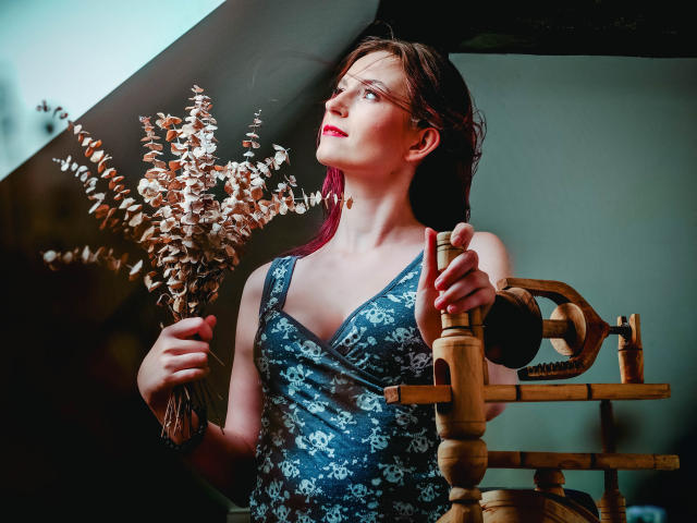 EmmaLust with red lipstick, holding dried flower bunch, wearing blue floral dress, standing by wooden chair, looking up, wet brown hair, hand on chair, soft light, pretty face.