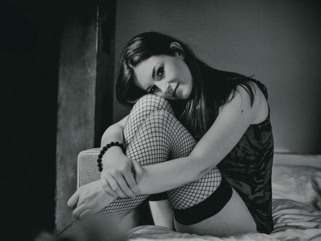 EmmaLust with dark hair, black fishnet stockings, and black lace top, hugging her knees on a bed, smiling slightly, wearing a black bead bracelet, in a black-and-white photo.