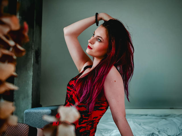 EmmaLust with red and purple hair, wearing a red and black tiger-print dress, sitting on a bed, arm raised, looking back, red lipstick, black bracelet, fishnet stockings.