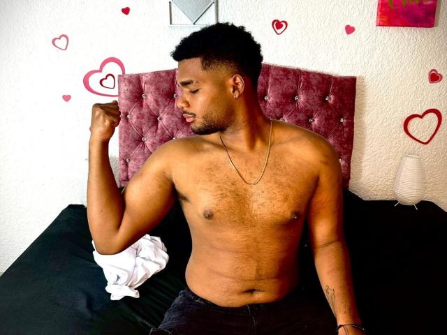 JacobSr, ohne Hemd, mit dunkler Haut, kurzen, lockigen Haaren und Bart, zeigt seinen rechten Arm, sitzt auf einem schwarzen Bett und trägt schwarze Hosen, mit einem pinkfarbenen, gepolsterten Kopfteil..