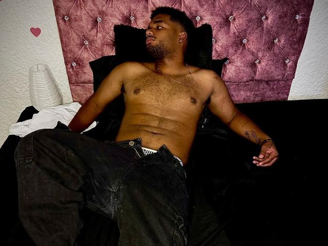 Shirtless JacobSr with a dark beard, chest hair, and a tattoo on his right arm, lying on a black bed, wearing unbuttoned black pants, with a pink tufted headboard.
