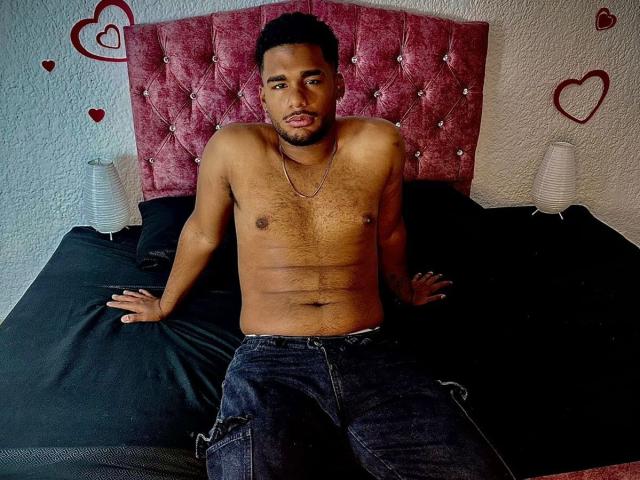 JacobSr ohne Hemd, mit behaarter Brust, dunkler Haut, kurzen schwarzen Haaren und einer Halskette, sitzt auf einem schwarzen Bett und trägt dunkle Jeans, mit weißen Lampen auf beiden Seiten..