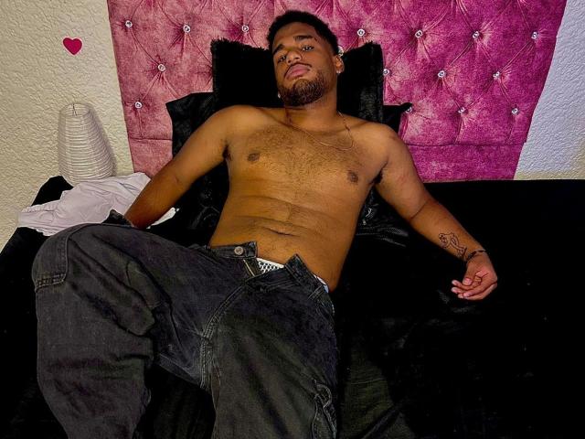 JacobSr ist ohne Hemd, liegt auf einem schwarzen Bett mit einem rosa, gepolsterten Kopfteil. Er trägt dunkle Jeans, hat einen Bart und ein Tattoo auf seinem rechten Arm. Seine Brust ist behaart..
