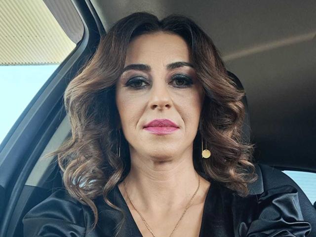 StellaDirty69X has wavy brown hair, dark makeup, pink lipstick, and a black shirt. She's wearing gold heart earrings and a thin gold necklace. She's in a car.