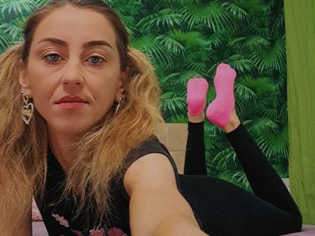 StellaDirty69X, blonde, lying on bed, black sleeveless shirt, pink socks, legs up, black leggings, green leafy wallpaper, wavy hair, blue eyes, slight smile, dangling earrings, selfie.