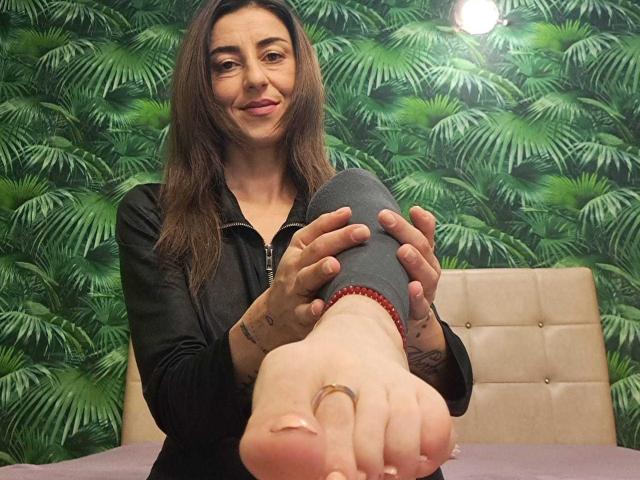 StellaDirty69X with long brown hair, black zip-up, holding her grey-sleeved foot up, showing her toes and gold ring, smiling, in a room with green leafy wallpaper.