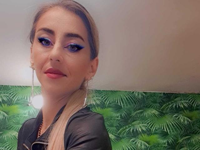 StellaDirty69X has blue eyeshadow, long blonde ponytail, black leather top, pink lipstick, and dangling earrings. She's smiling with a cute, confident look.