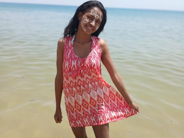 AmandaSexySexy, dark-skinned, wearing a pink and white diamond-patterned dress, has white face paint, smiling, standing in shallow blue-green water, pulling dress to the side.