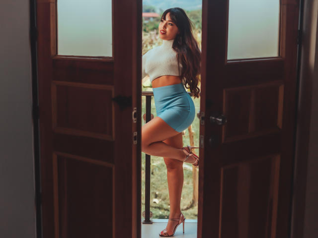 Heyly si trova in una porta, sexy in un crop top bianco e minigonna blu, mostrando il suo culo rotondo e tacchi a righe.