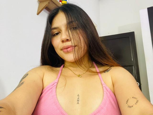 SophiaMarx with long brown hair, pink ribbed top, tongue out, tattoos on arms, chest, and collarbone, wearing a gold necklace, smiling at the camera.
