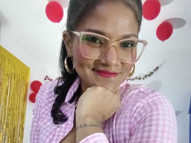 AlejandraBoobs with glasses, pink checkered shirt, and braid, smiling with pink lipstick, gold hoop earrings, and tattoo on wrist, holding cheek with hand. Red balloons in background.