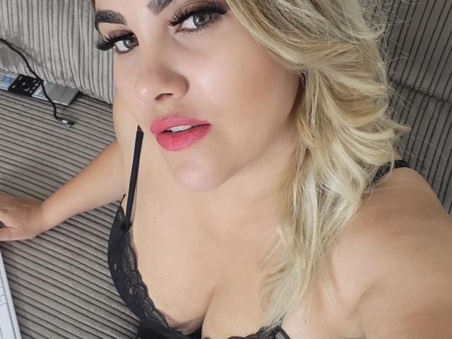 BabsTease with blonde hair, black lace bra, pink lipstick, and heavy eyeliner, looking up with a slight smile, showing her tits and a bit of her cleavage.