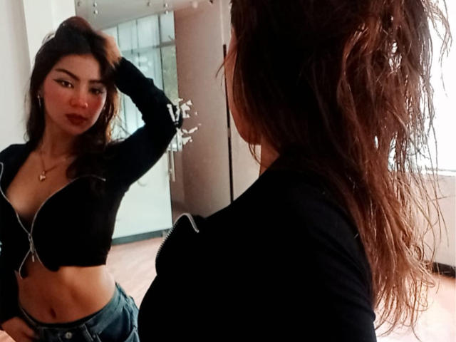 AshleyMiler with wet hair, black zip-up, and blue jeans, showing her tits in a mirror, hand in hair, looking at reflection, in a dimly lit room.