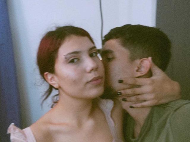 SpecialFriendsBenefits with red hair and black nails has a pensive look while a dark-haired guy with black nails kisses her neck. She's wearing a pink top, he's in green.