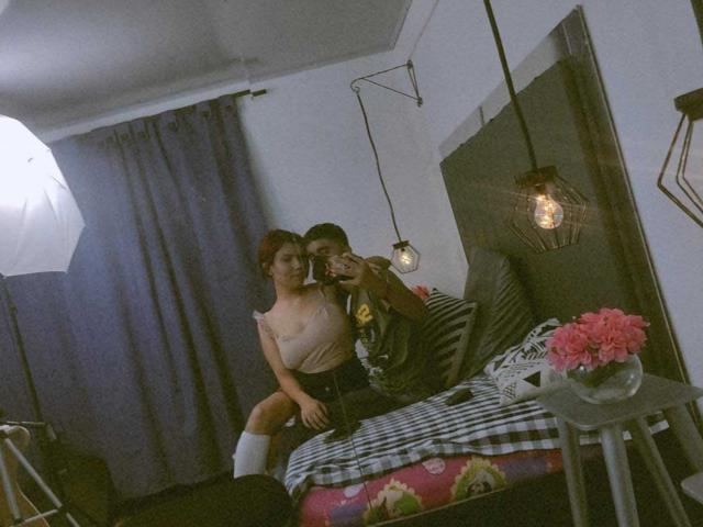 SpecialFriendsBenefits with small tits and a light pink top, sitting on a bed, holding a phone. SpecialFriendsBenefits with a dark shirt, taking the photo. Pink flowers on a table.