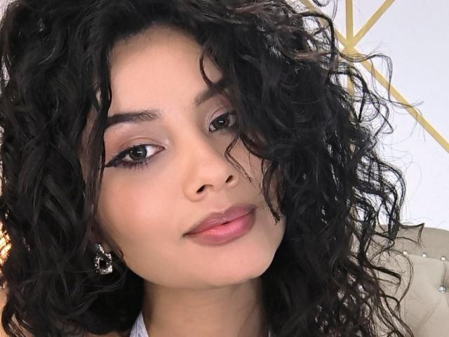 CanelaLebrad's face with curly black hair, brown eyes, and pink lipstick. She's wearing silver earrings and has a slight smile. Her makeup includes winged eyeliner.