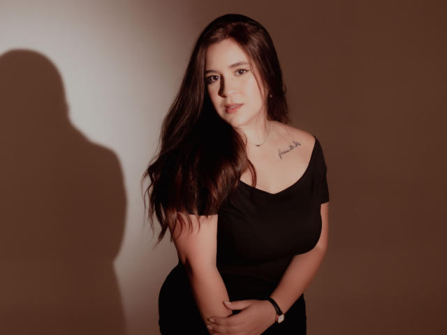 EmmyGray with long brown hair, black off-shoulder top, and tattoo on right chest, arms crossed, shadow on wall, slight smile, soft brown lighting, standing.