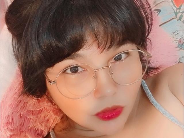 PrittyAlicia with round glasses, pink lipstick, and short black hair, lying on a pink fluffy pillow, wearing a gray strap top, looking directly at the camera.