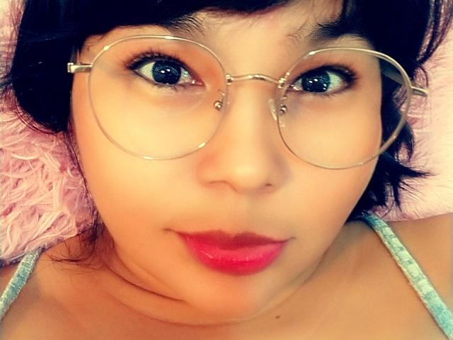 PrittyAlicia with big blue eyes, wearing round glasses, has pink lipstick, and short black hair, showing a cute, slightly chubby face with a subtle smile.