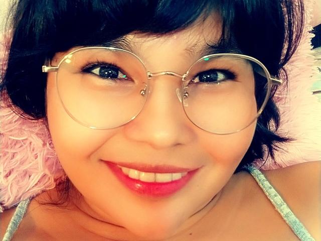 PrittyAlicia with shiny round glasses, dark short hair, and pink lipstick smiling at the camera. Her blue eyes and cute face are visible. She's wearing a gray top.