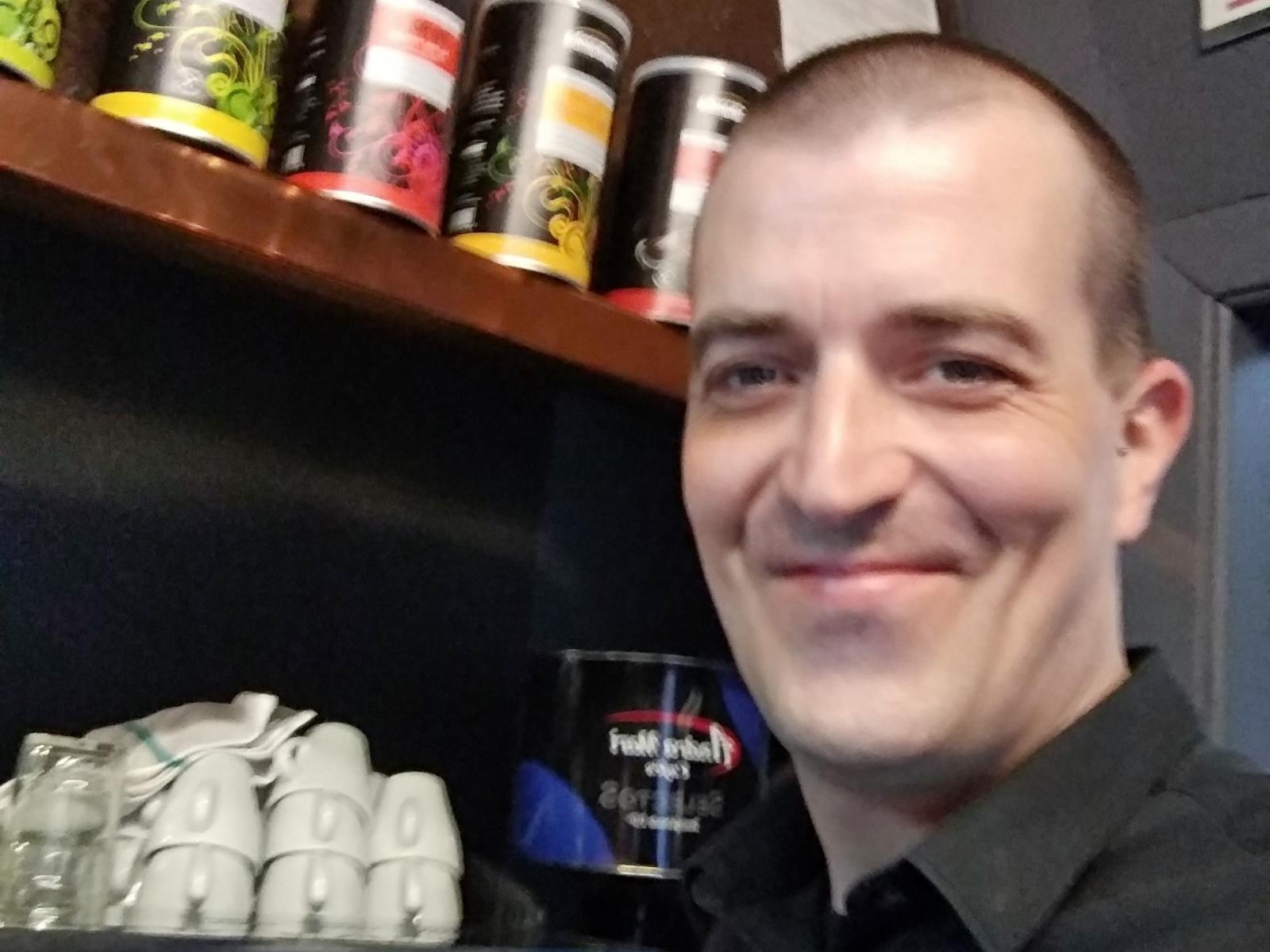 Andres Alan, calvo con un leggero sorriso, indossa una camicia scura e si trova di fronte a uno scaffale con lattine e una tazza bianca; ha il viso rasato..